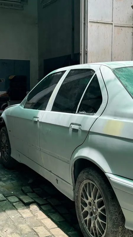 White-primed E36-type sedan side with BBS-style wheel in the workshop yard