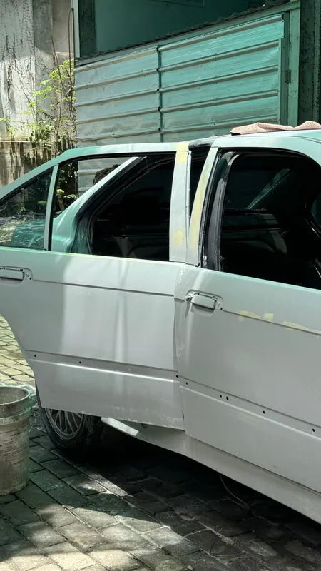 White-primed E36 with the door open and the cabin stripped, in front of the paint-bay wall