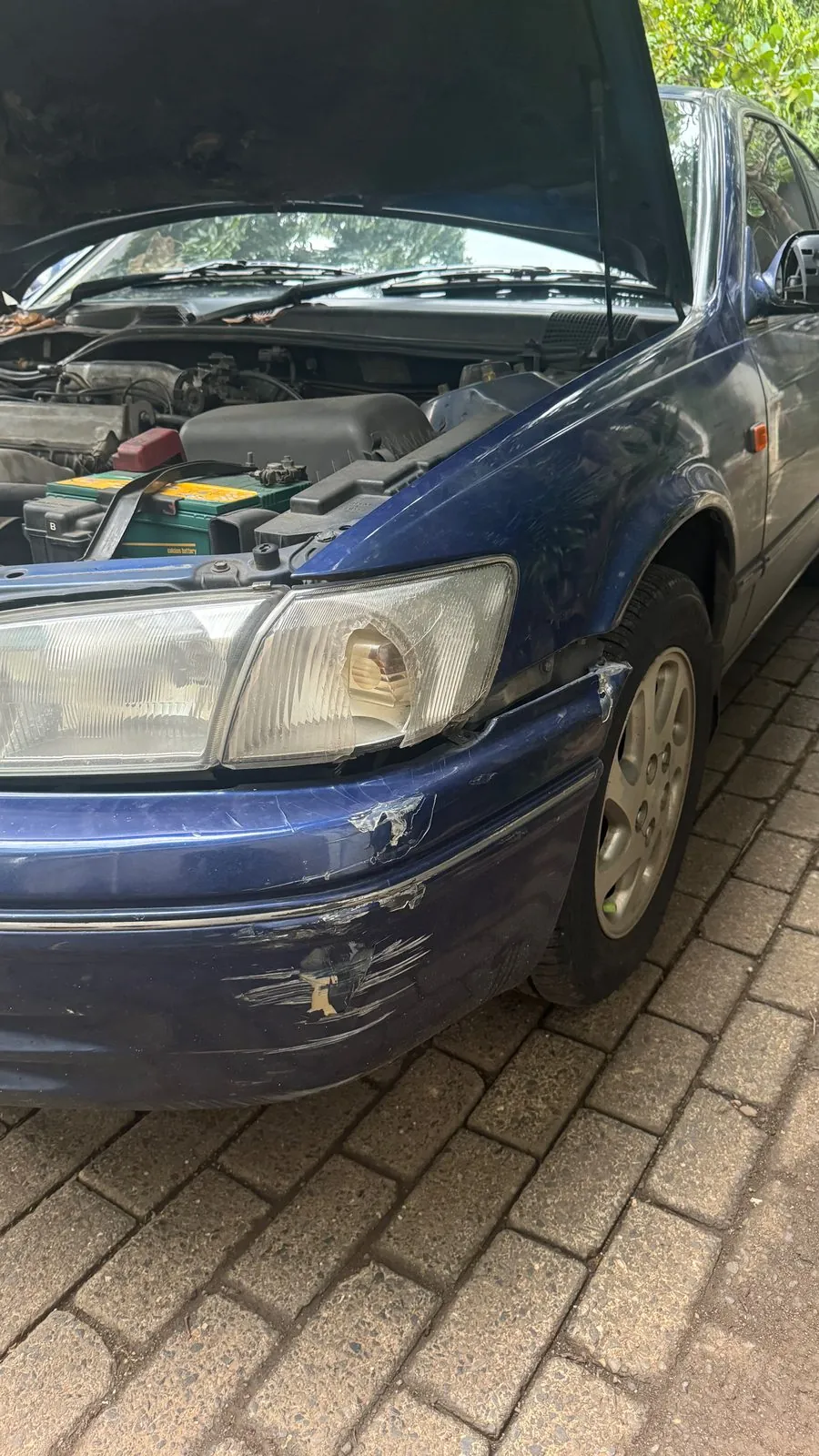 Front corner of the blue Camry with a scraped bumper edge and bonnet up